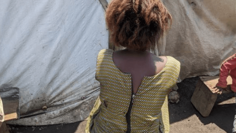 A woman with reddish-brown hair wearing a yellow and black patterned sleeveless shirt stands with her back to the camera, facing a makeshift shelter made of white plastic sheeting.