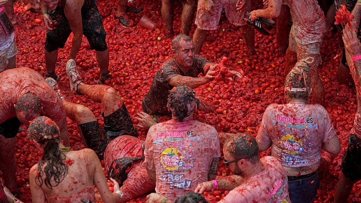 Hundreds of people covered in red tomato pulp participate in a massive food fight, many wearing shirts with festival logos and covered head to toe in tomato residue.