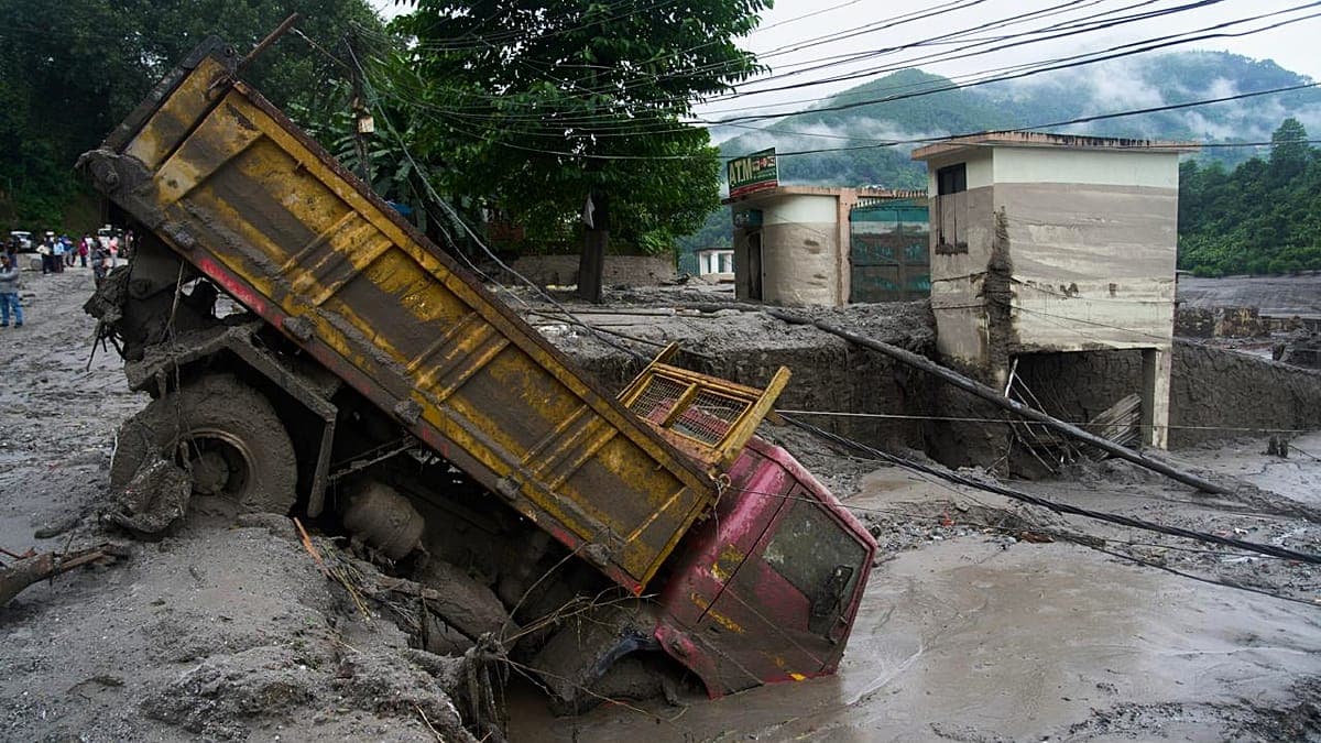 Sebuah truk berwarna kuning dan merah yang terperosok dalam lumpur dan air, dengan bak muatan terbuka, di area yang dipenuhi material tanah liat keabu-abuan di samping bangunan-bangunan sederhana dan pegunungan yang tertutup pohon di latar belakang.