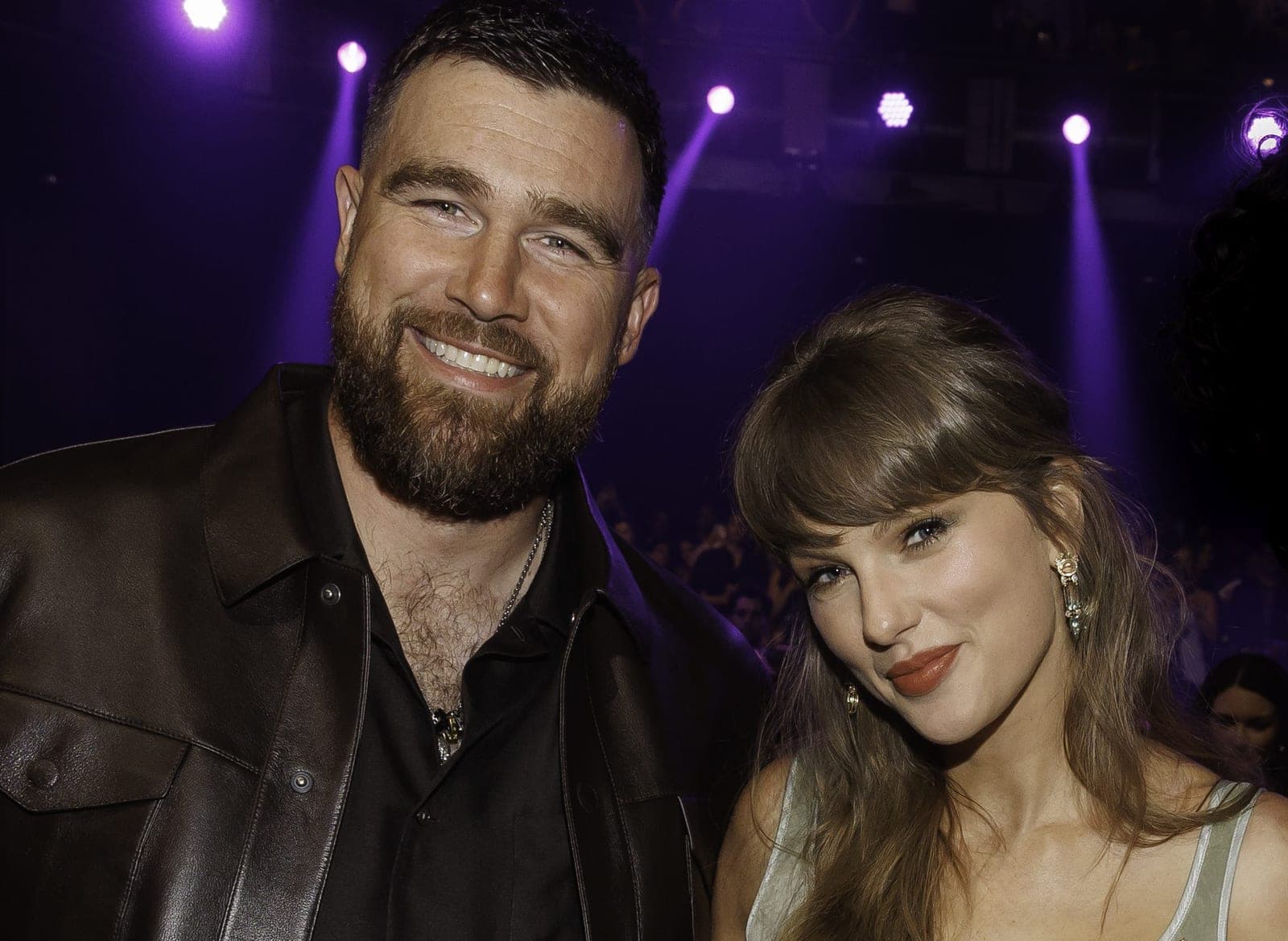 A bearded man in a black leather jacket and a woman with long brown hair and bangs stand together smiling at what appears to be an indoor event with purple lighting in the background.