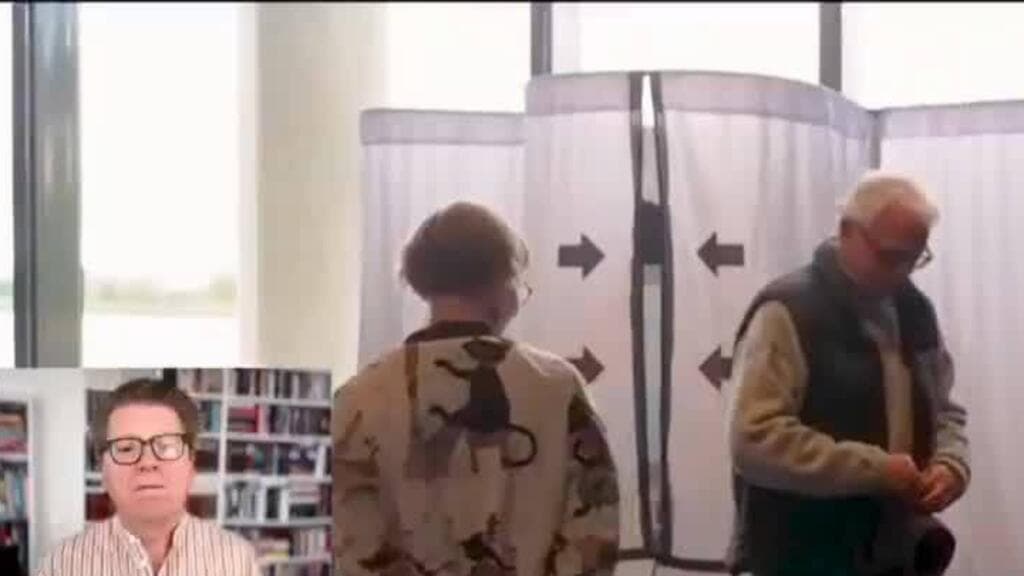 Two people stand near a voting booth with directional arrows inside a bright room with bookshelves visible.