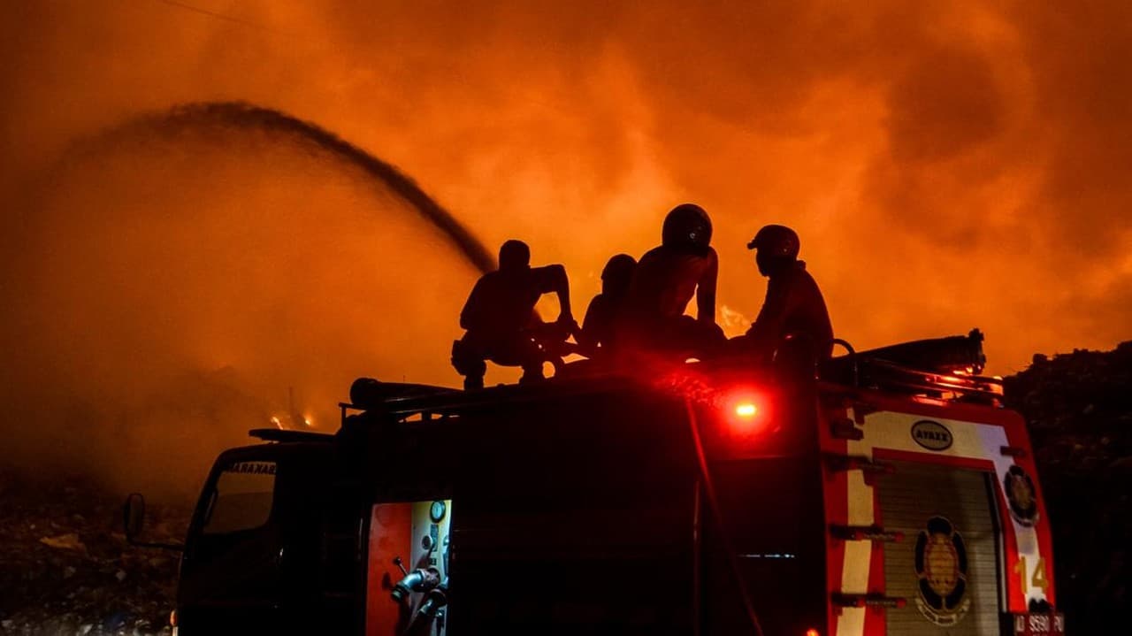 Petugas pemadam kebakaran bersiap menyiram api dengan selang air dari atas kendaraan pemadam kebakaran, dengan latar belakang asap dan api yang membara berwarna oranye terang.