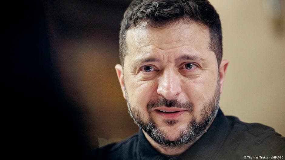 A man with dark hair and facial hair wearing a dark shirt speaks with a serious expression.