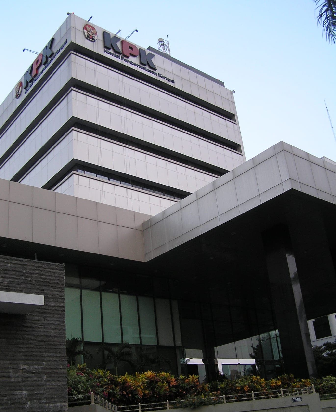 Gedung bertingkat dengan signage KPK di bagian atas, memiliki arsitektur modern dengan panel putih dan jendela horizontal besar, serta area ground floor dengan kaca gelap dan taman di depannya.