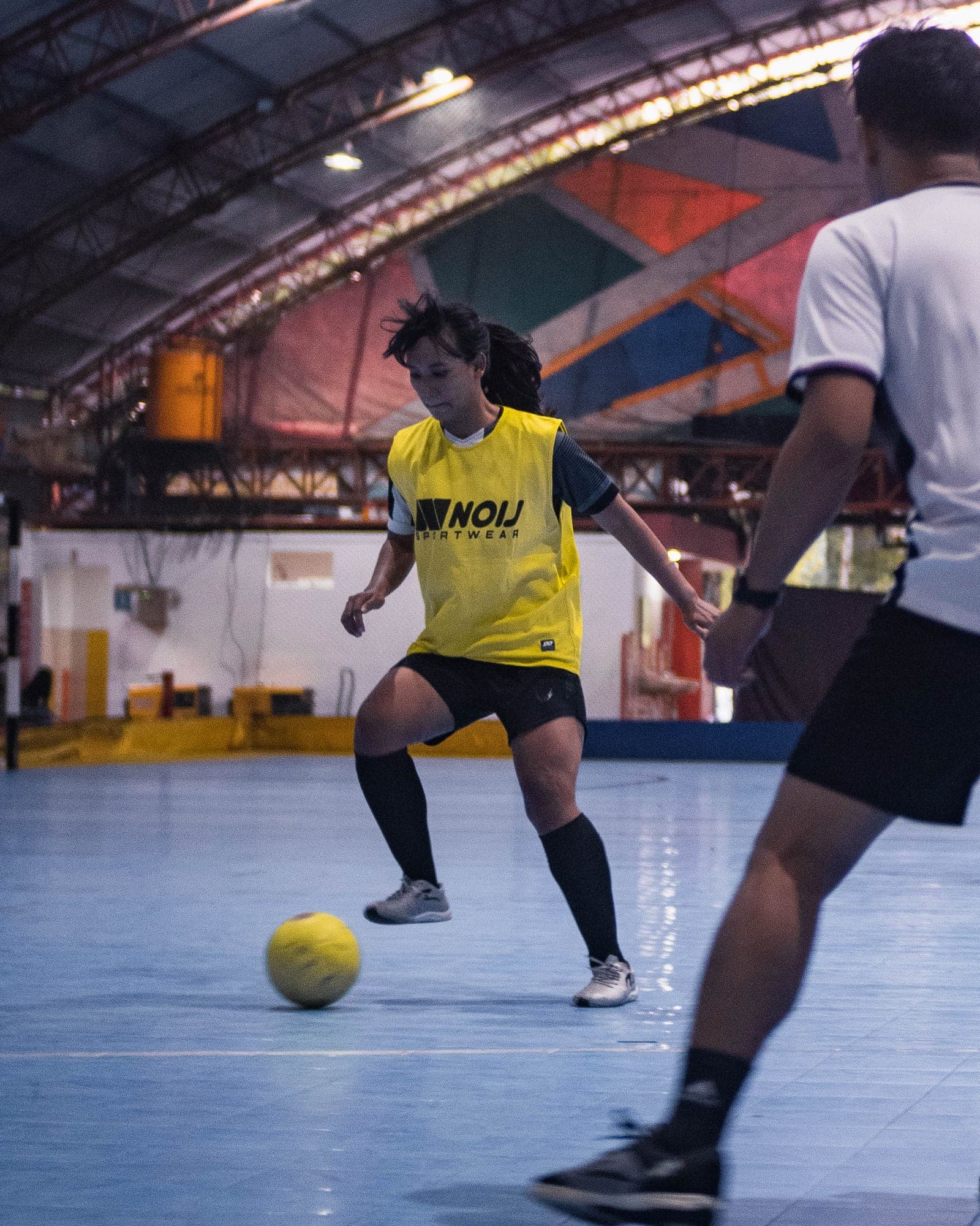 Seorang pemain futsal perempuan mengenakan seragam kuning dengan logo "KNOLL FOOTWEAR" sedang menggiring bola kuning di dalam lapangan futsal bercetak biru, dengan pemain lain mengenakan seragam putih terlihat di sebelah kanan.