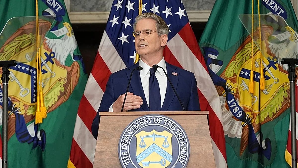 A man in a dark suit and tie stands at a Department of the Treasury podium, speaking with the U.S. flag and two green state flags visible behind him.