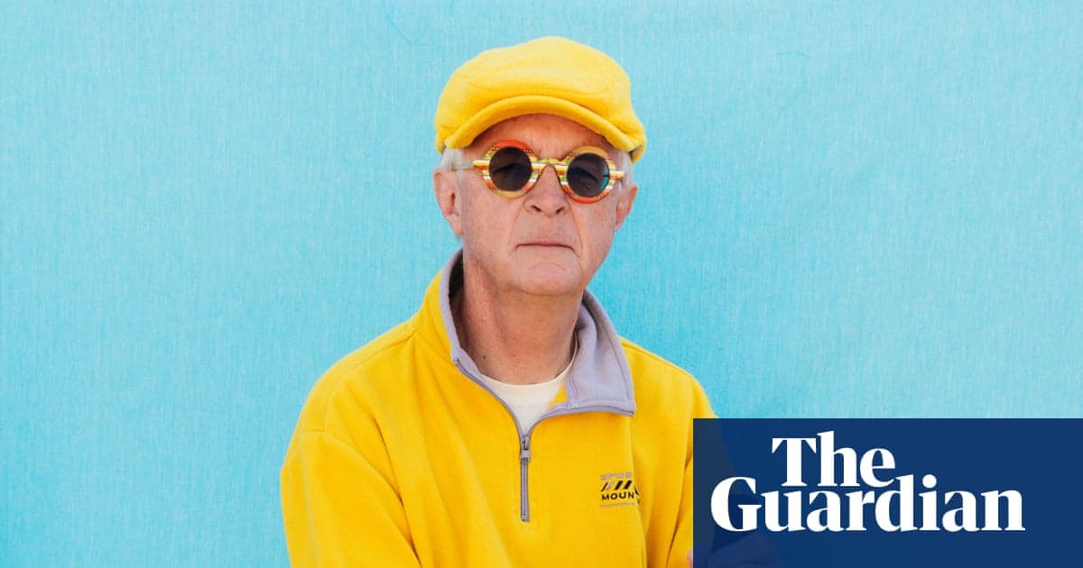 A person wearing a bright yellow cap, round-framed sunglasses with orange-tinted lenses, and a yellow zip-up jacket over a lavender collar, posed against a turquoise background.