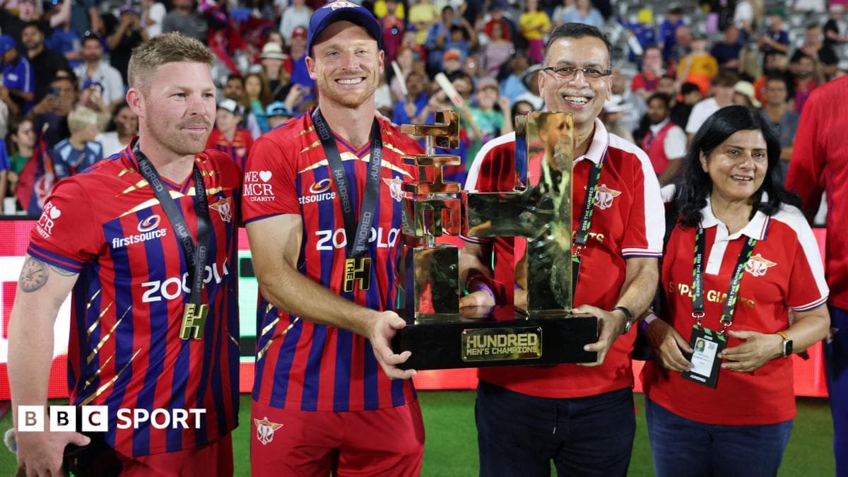 Players and staff members in red and blue striped cricket uniforms hold a large trophy on a cricket field in front of a crowded stadium.