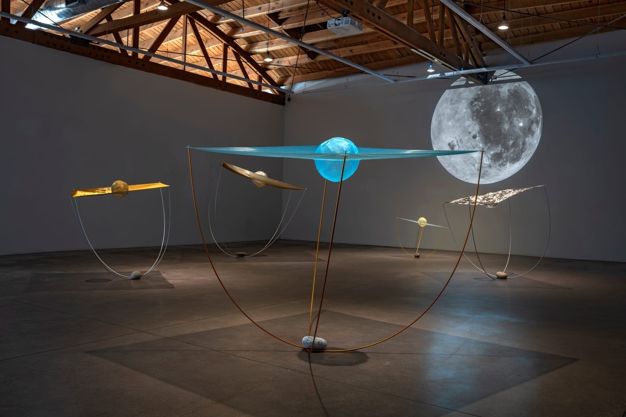 An art installation in a gallery space featuring suspended sculptural elements including a large illuminated blue sphere, a detailed photographic moon, and geometric wire and metal forms with golden and metallic spheres at various points.