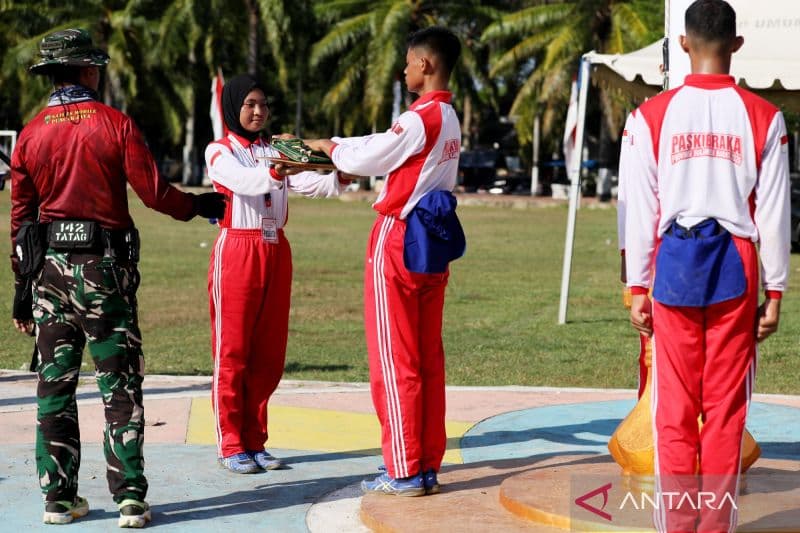 Sejumlah anggota tim berkostum merah putih sedang berbaris dan berinteraksi di sebuah lapangan berumput dengan latar belakang pohon kelapa.