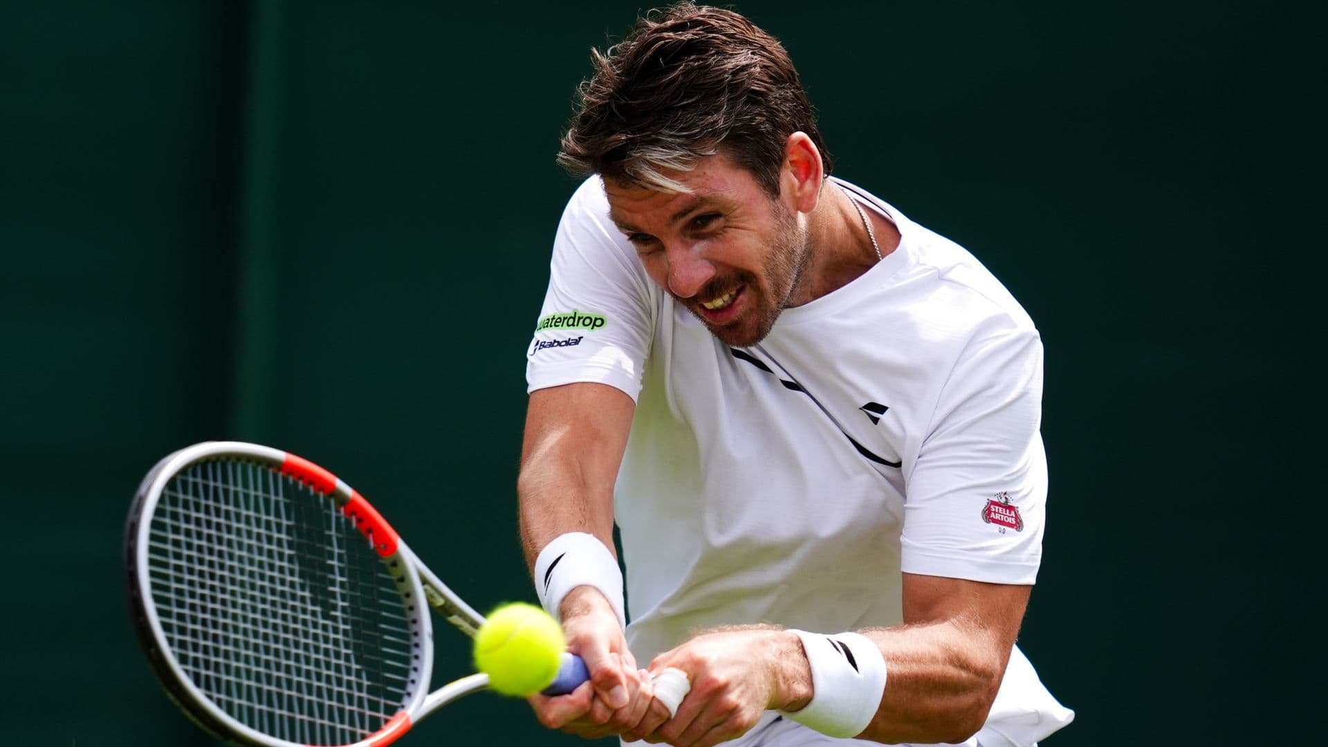 A male tennis player in white athletic wear is captured mid-swing during a tennis match, holding a red-trimmed racket with a yellow tennis ball in frame.