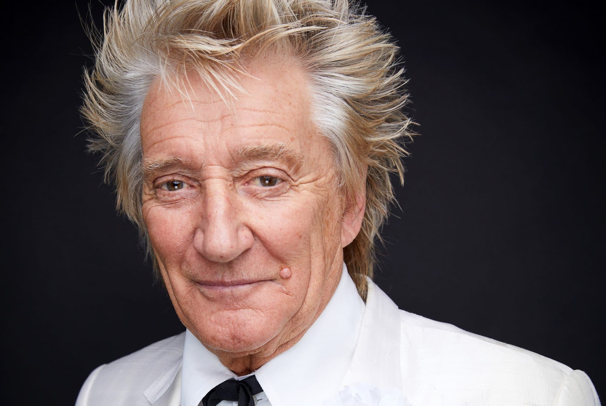 An older man with shoulder-length white and blonde hair wearing a white blazer and black tie against a dark background.