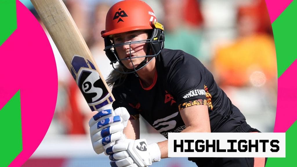 A female cricket batter wearing a red helmet and black uniform holds a cricket bat in a ready position during a match.