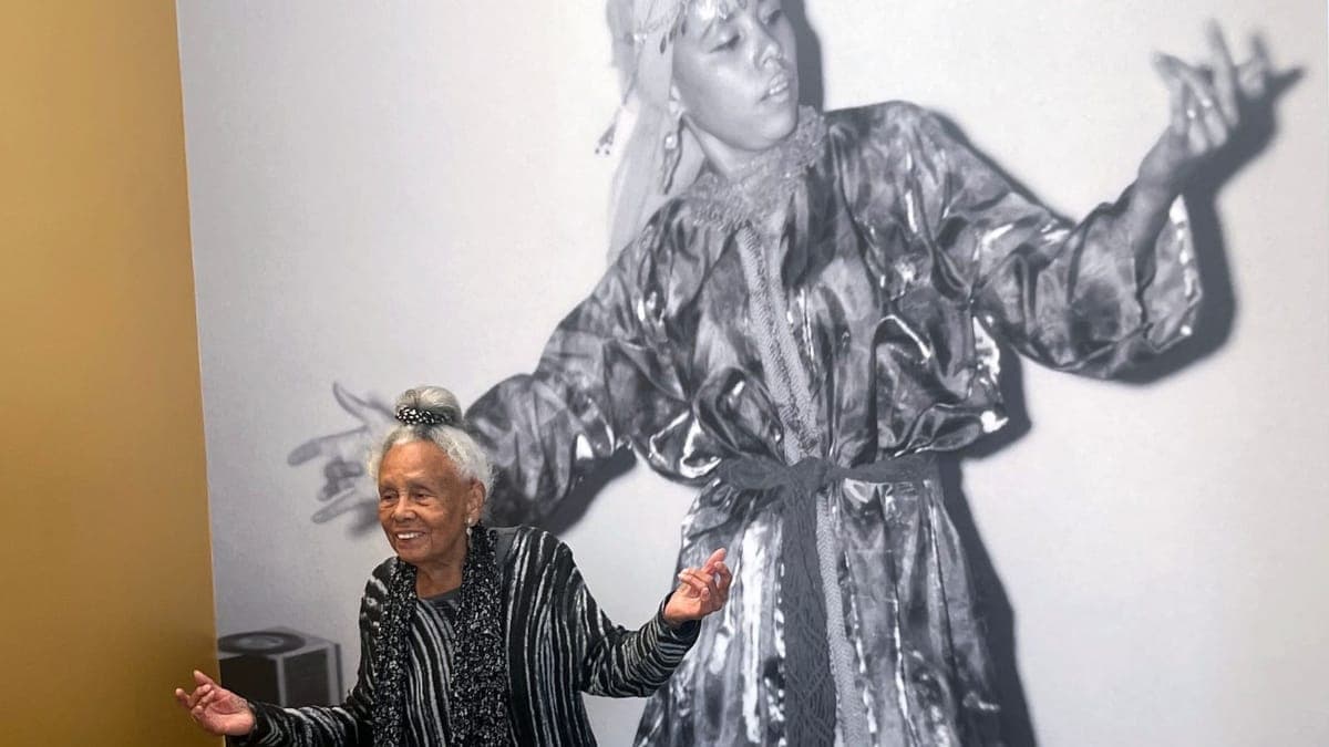 An elderly woman with white hair stands in front of a large black and white photograph of a figure in an ornate robe with arms outstretched.