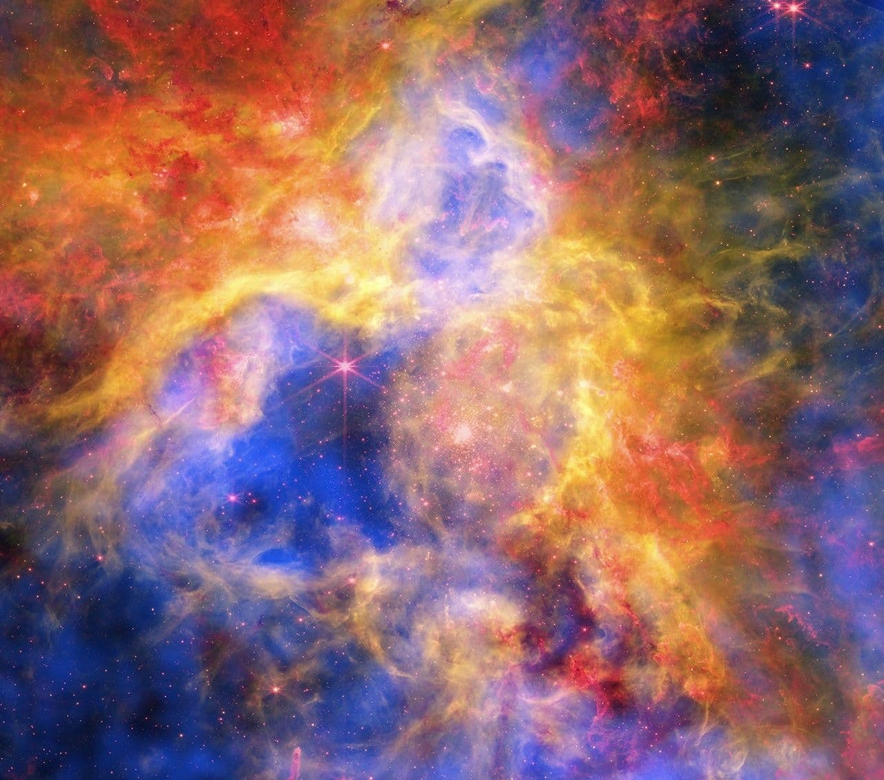 A colorful composite image of the Tarantula Nebula showing swirling clouds of gas and dust in shades of blue, yellow, orange, and red, with bright stars scattered throughout.
