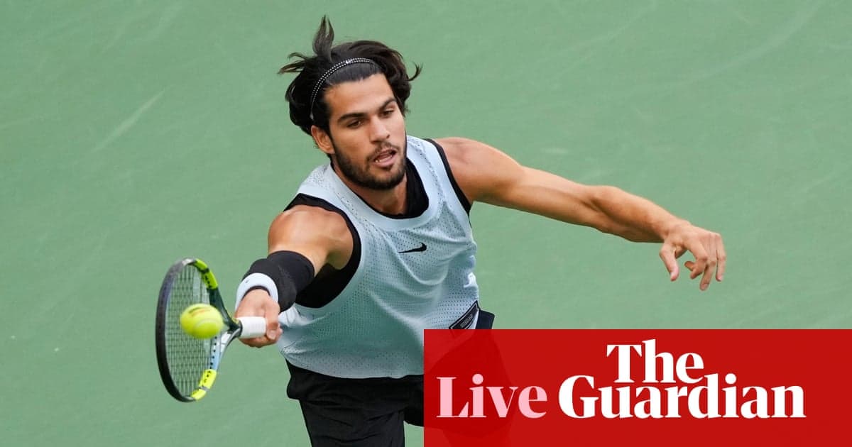 A male tennis player in a light blue sleeveless shirt hits a forehand shot during a match on a green hard court.