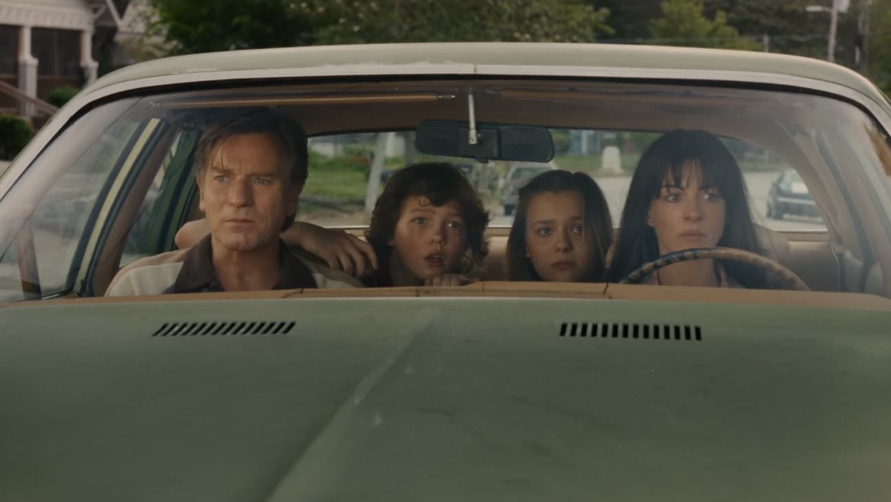 Four people sit inside a light-colored sedan, looking ahead with expressions of concern or worry.