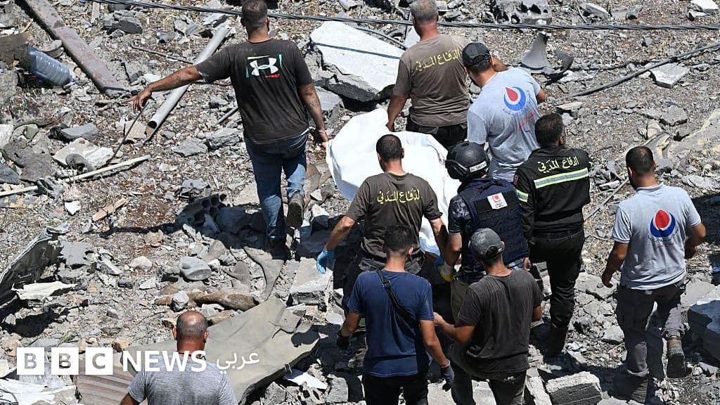 Emergency responders and civilians work among concrete rubble and debris to remove a body wrapped in white cloth from a destroyed building.
