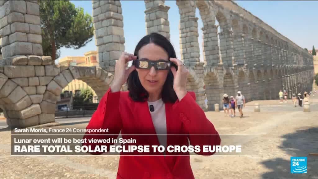 A female journalist in a red blazer stands before an ancient Roman aqueduct with multiple stone arches, adjusting her sunglasses while speaking to camera.
