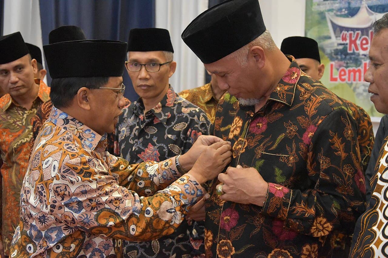 Para pejabat pemerintah dan akademisi berbatik tradisional dan peci hitam berdiskusi dalam acara resmi di ruangan berdekorasi.