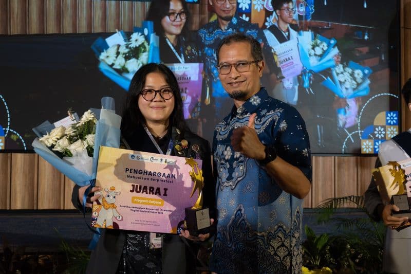 Seorang mahasiswi memakai kacamata sedang berdiri di samping seorang pria yang mengenakan baju batik biru tua, keduanya tersenyum sambil memegang piala dan buket bunga putih di atas panggung, dengan layar besar menampilkan foto-foto penerima penghargaan lainnya di latar belakang.