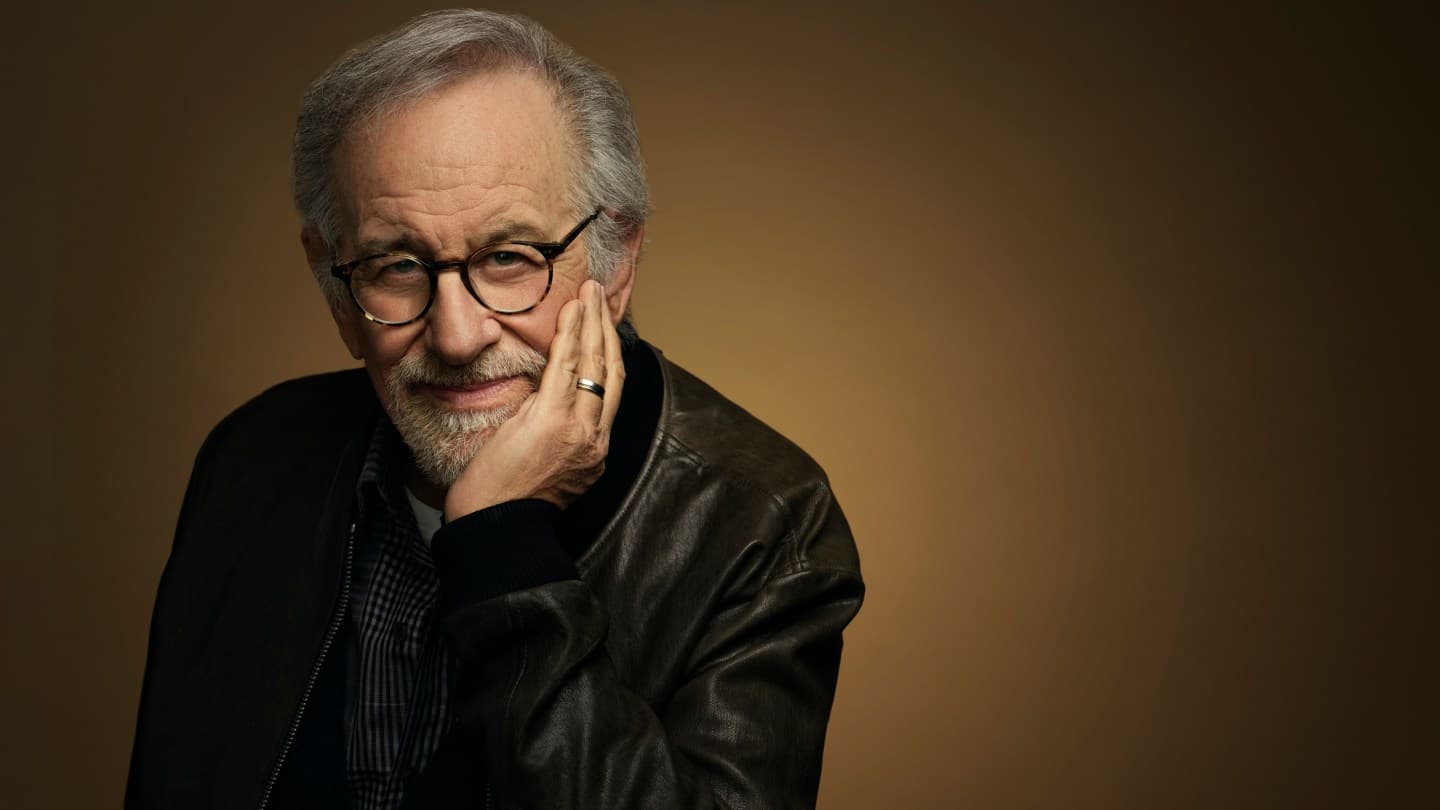 A man with gray hair and beard wearing glasses and a black leather jacket over a dark plaid shirt, resting his chin on his hand against a warm brown background.