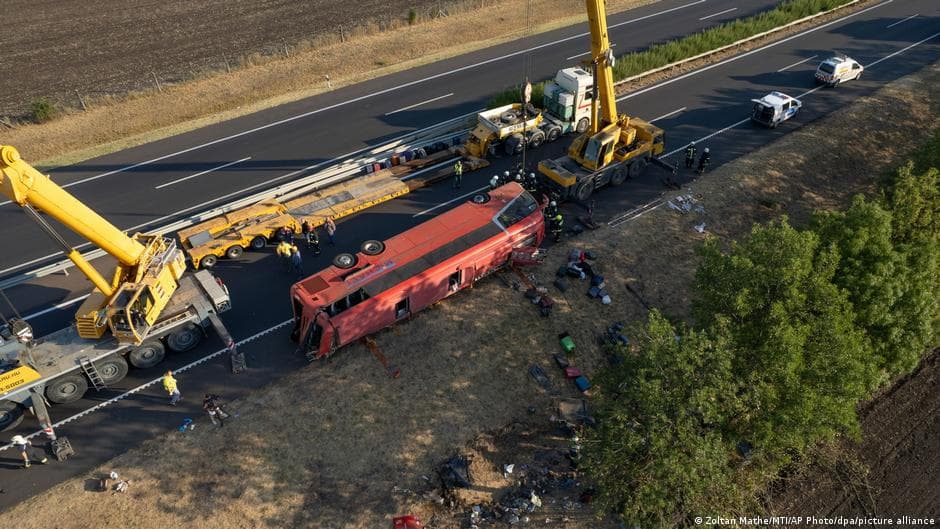 Aerial view of a red coach overturned on the side of a highway with heavy recovery equipment and emergency responders gathered around the wreckage, with police vehicles visible on the road.