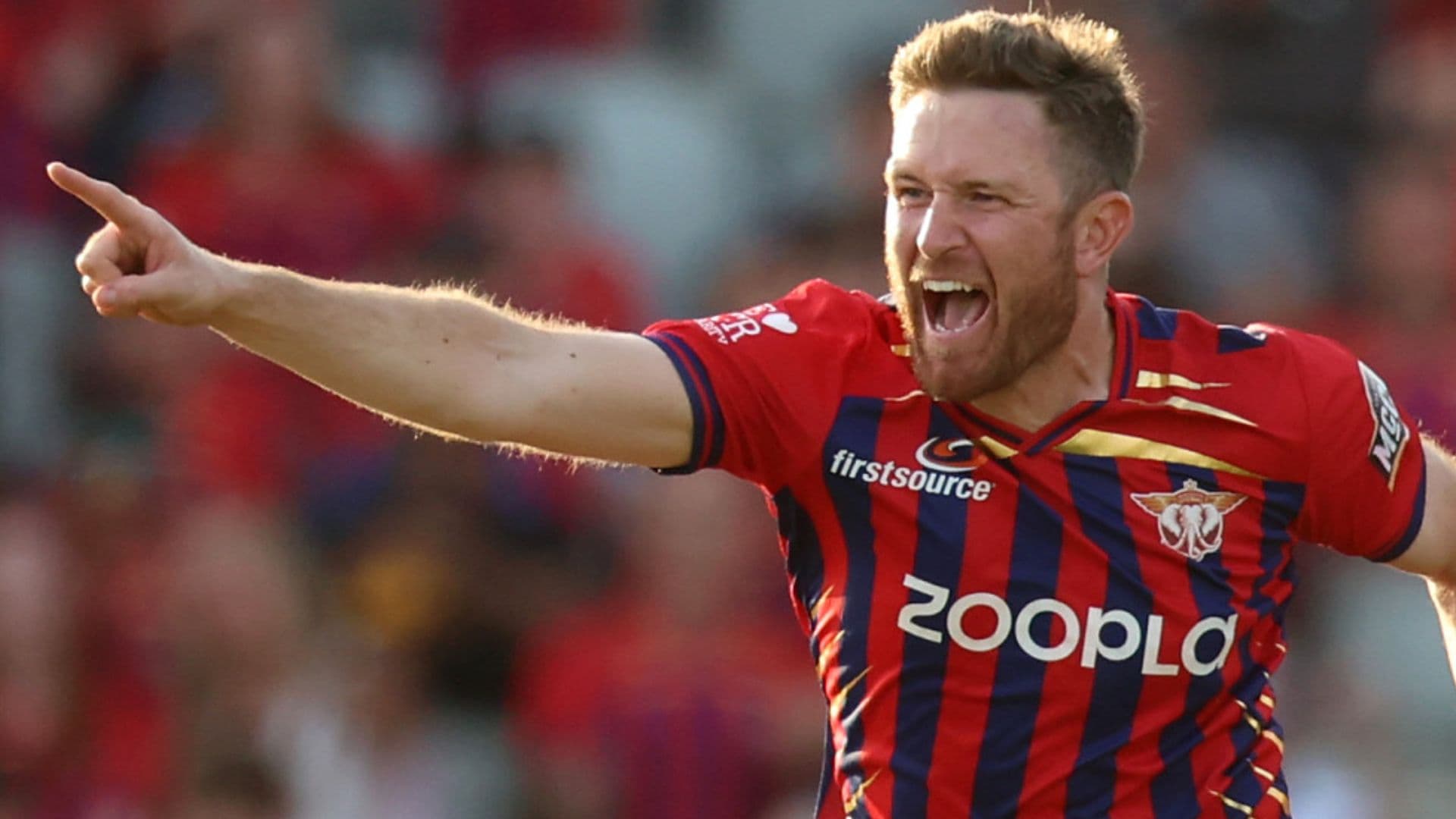A cricket player in a red and navy striped jersey celebrates with a pointing gesture and open-mouthed expression during a match.