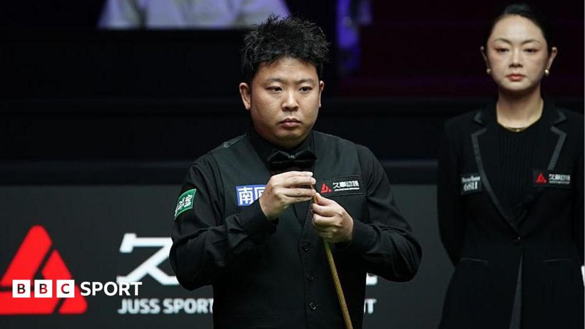 A snooker player in a black waistcoat holds a cue during a match, with another person in black formal wear standing beside him.