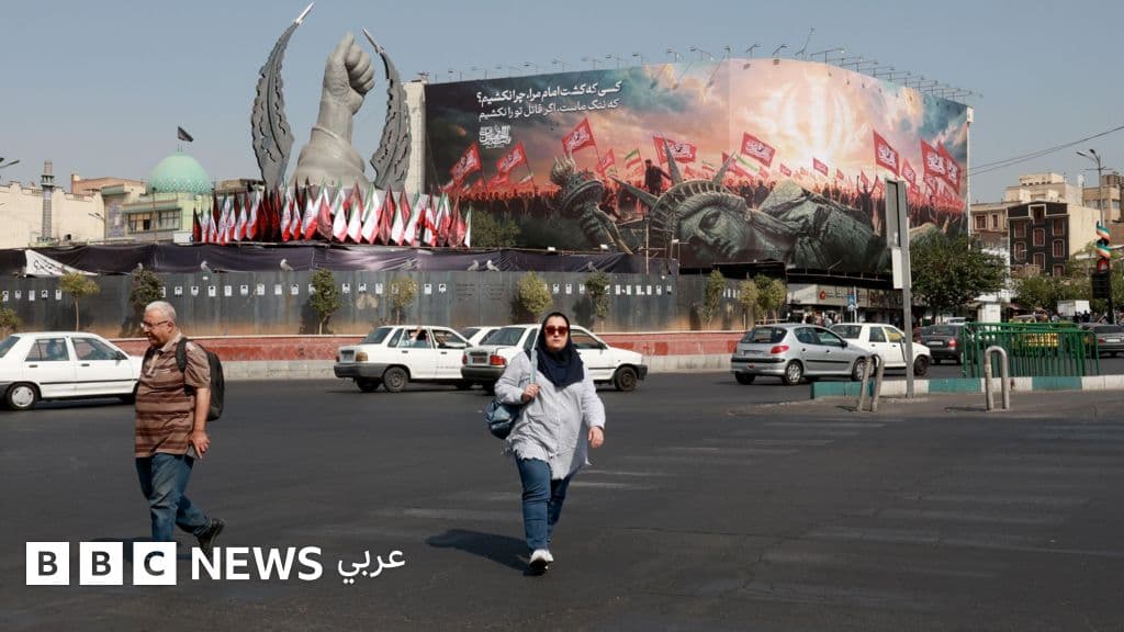 A public square in Iran featuring a large monument with white and red flags, alongside a billboard with red flags and Arabic text, with pedestrians and parked cars visible in the foreground.