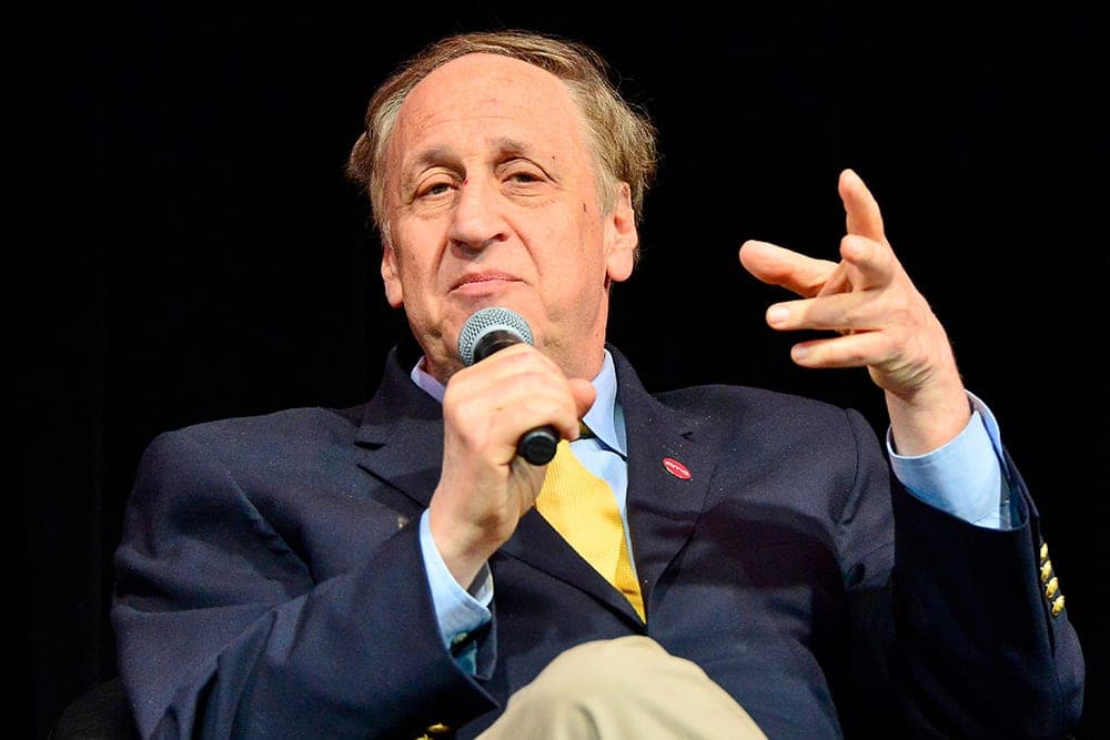 A man in a dark suit with a light blue shirt and yellow tie holds a microphone and gestures with his right hand while speaking.