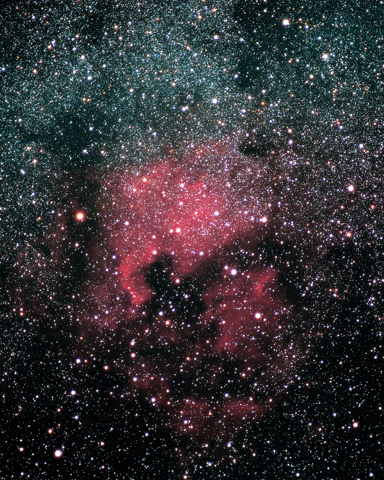 A deep space photograph showing a dense star field with a prominent nebula in the center displaying red and pink ionized hydrogen gas, surrounded by thousands of stars of varying brightness.