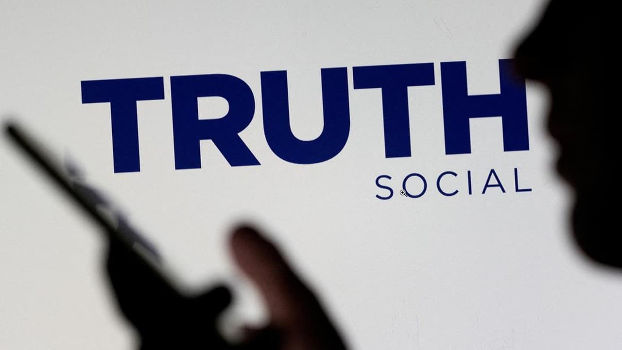 A hand pointing at a smartphone screen displaying the "TRUTH Social" logo in dark blue letters on a white background.