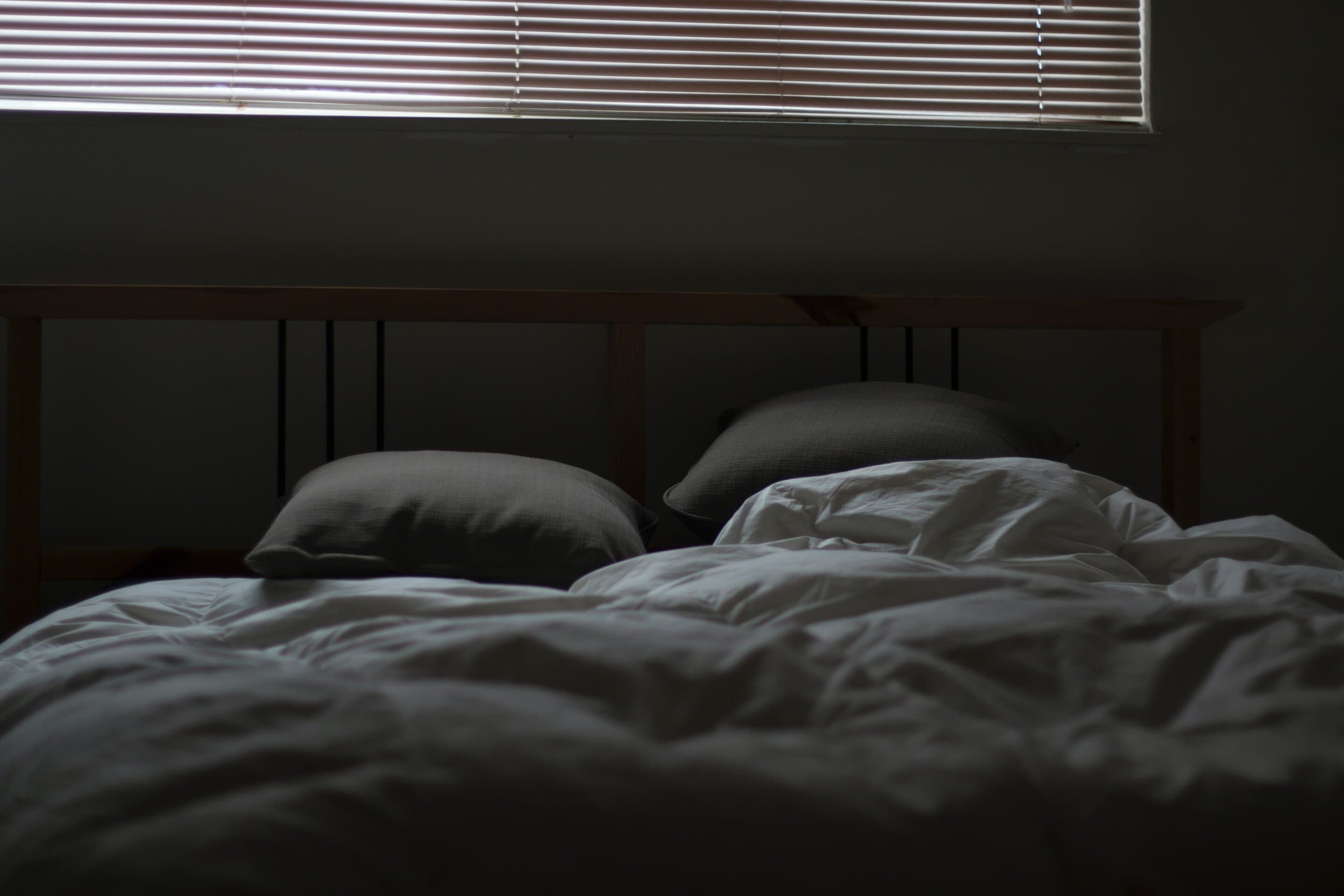 An unmade bed with gray and white bedding and dark pillows against a dark headboard, with horizontal blinds visible in a window above.