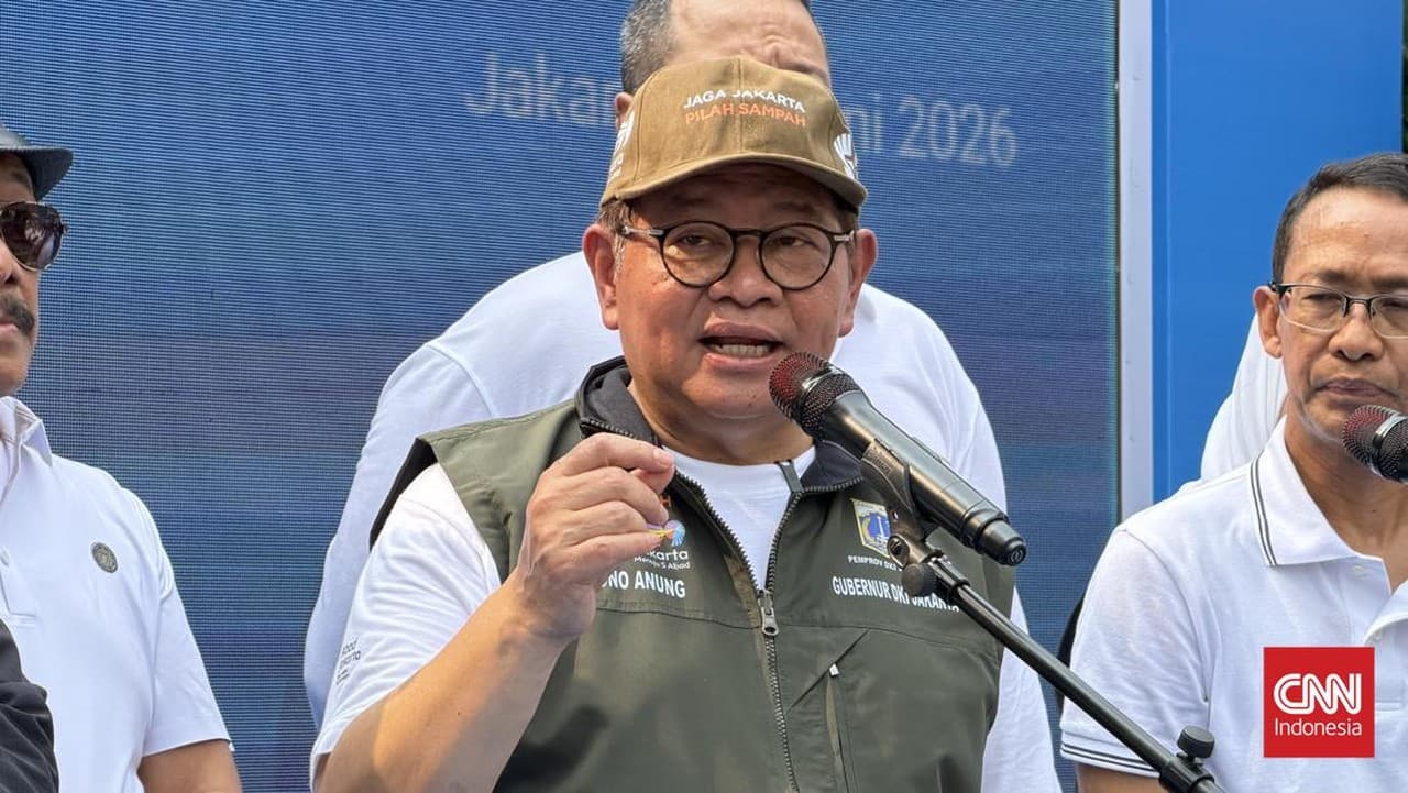 Seorang pria dengan kacamata berbicara di depan mikrofon sambil mengenakan topi dan rompi hijau dengan tulisan tentang Jakarta.
