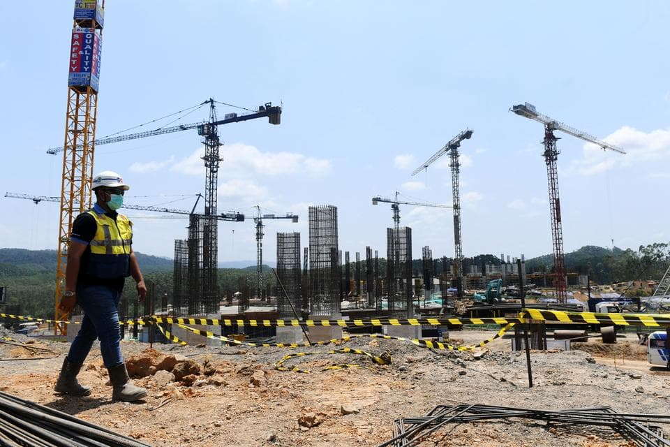 Seorang pekerja konstruksi mengenakan helm dan rompi keselamatan kuning berdiri di lokasi proyek bangunan dengan beberapa crane dan struktur baja yang sedang dibangun.