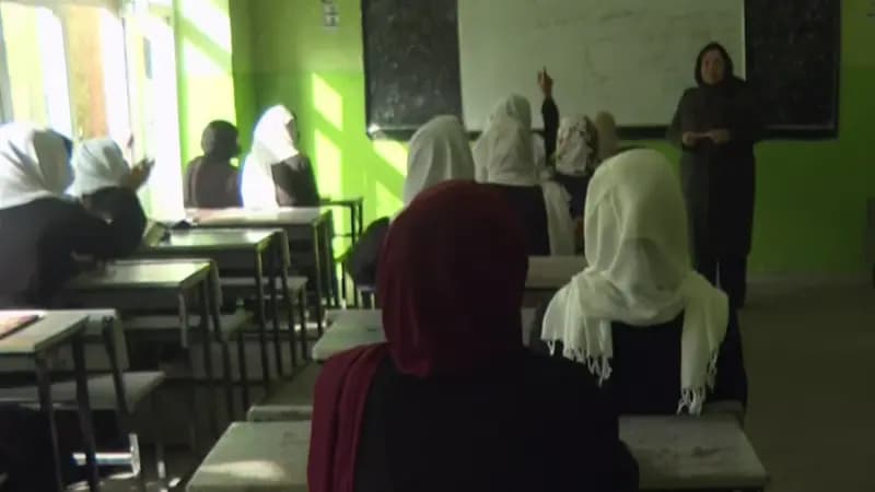 A classroom with bright green walls where students wearing headscarves sit at desks while a teacher stands at the front of the room.