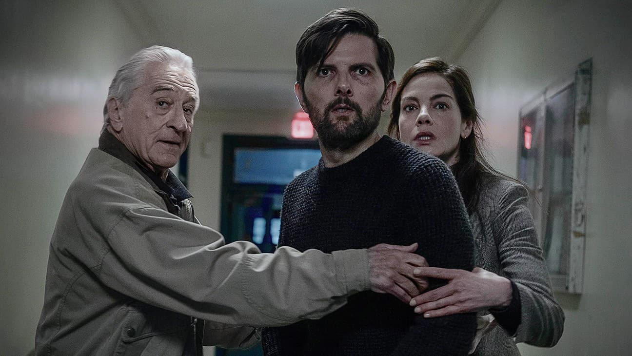 Three actors stand together in an indoor hallway setting, looking toward the camera with serious expressions.