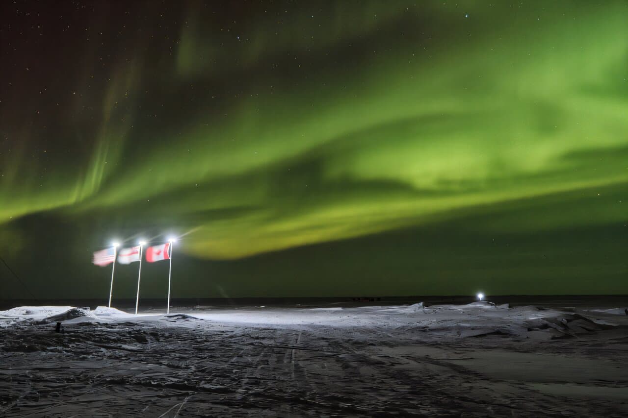 Three illuminated flags planted in snow with the aurora borealis glowing green above them at night.