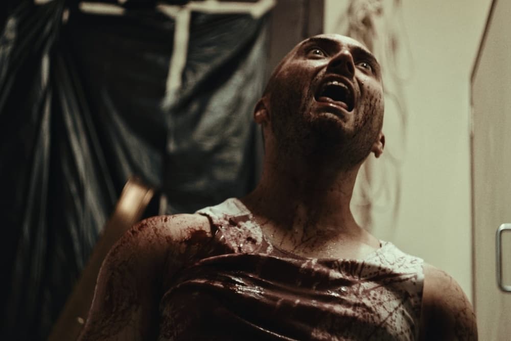 A man with a distressed expression looks upward with his mouth open, appearing frightened or in pain, with visible dirt or debris on his bare chest and shoulders.