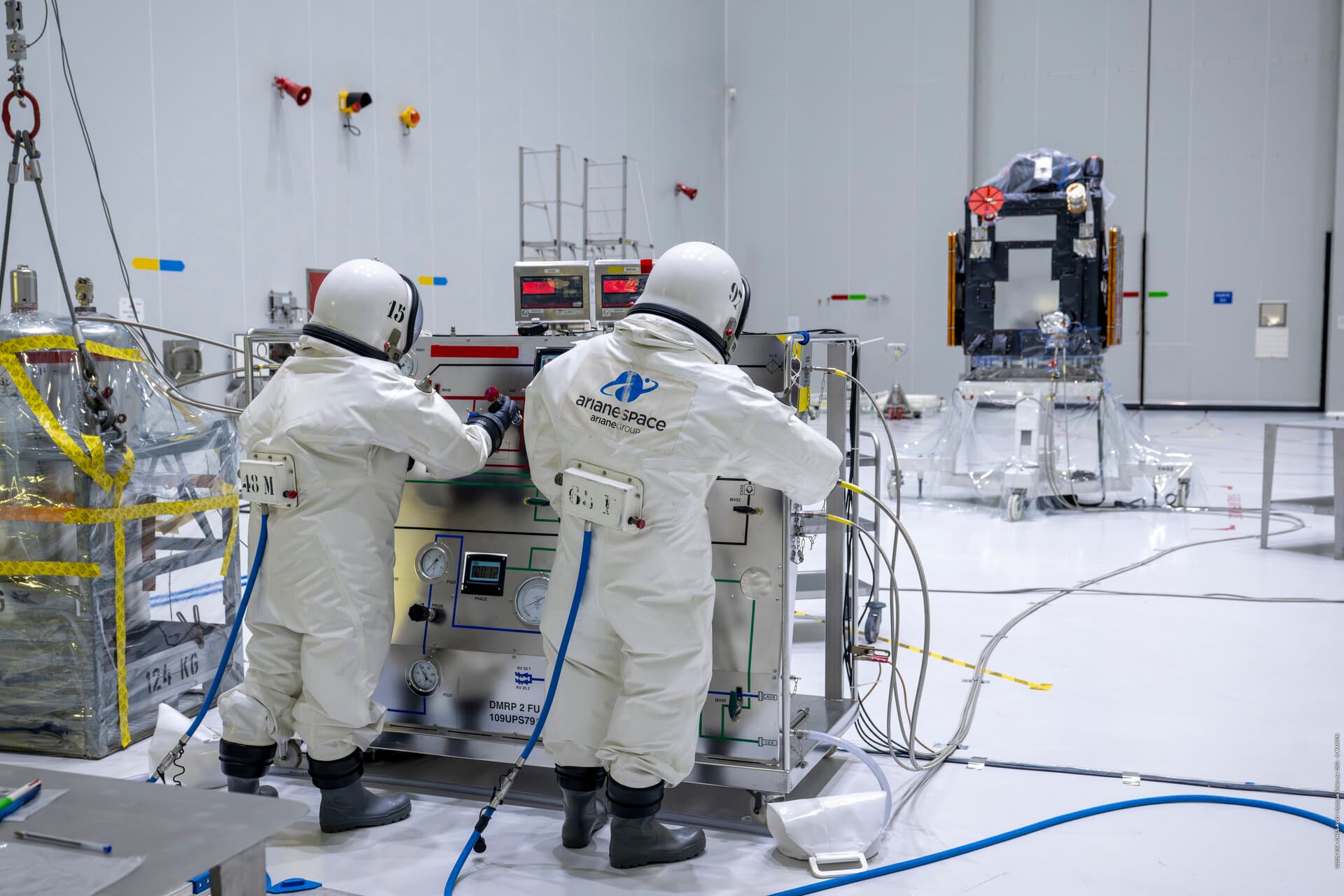 Two technicians in white spacesuits and helmets work on a satellite component inside a clean laboratory facility, with various equipment and monitoring instruments visible in the background.