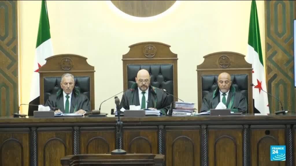 Three judges in dark robes and green sashes sit behind a wooden bench in a courtroom, with Syrian flags and official emblems visible in the background.