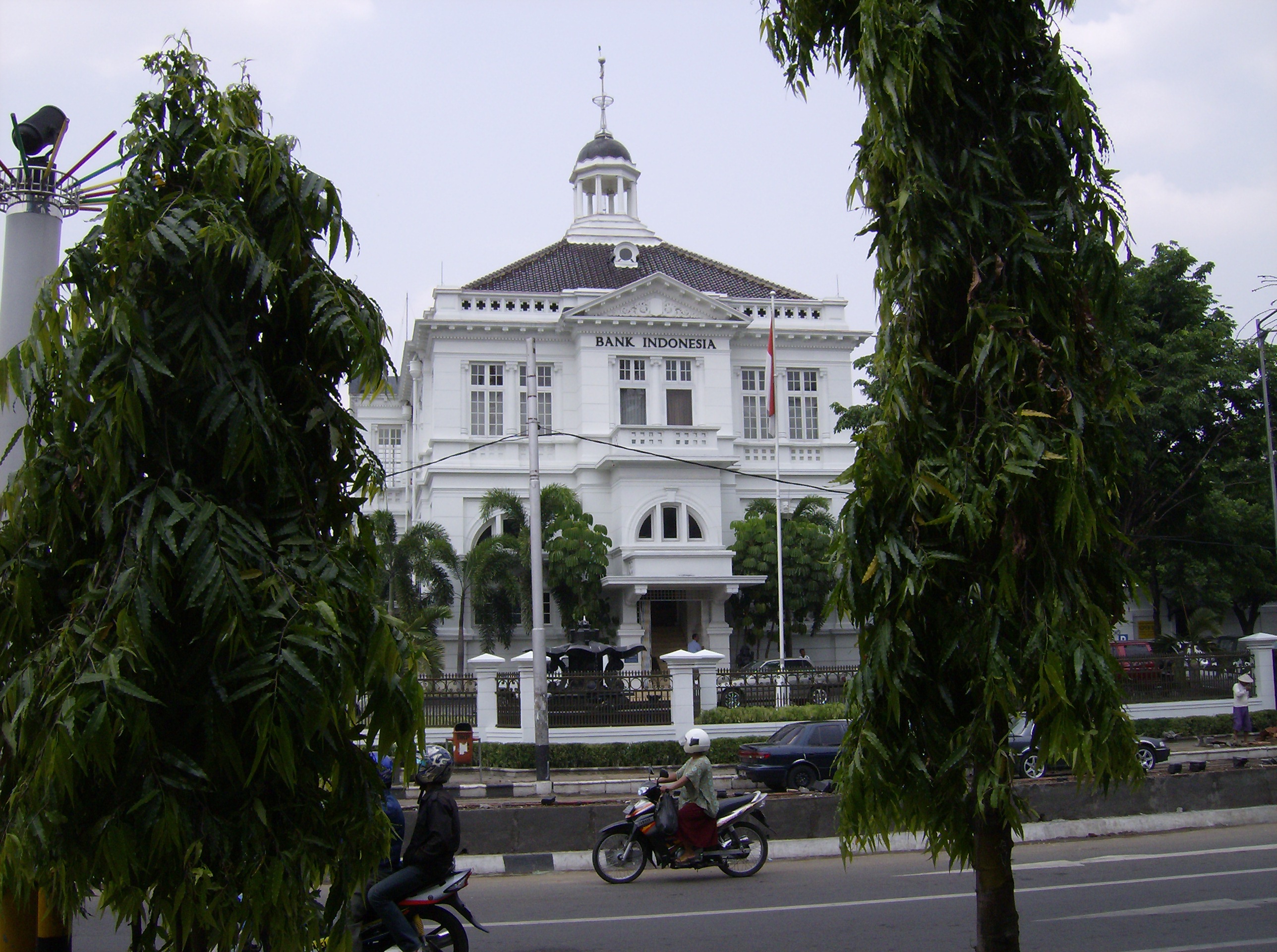 Gedung putih berarsitektur kolonial dengan kubah berlapis emas di puncaknya dan tulisan "BANK INDONESIA" di fasadnya, dikelilingi pohon-pohon hijau dan lalu lintas jalan raya.