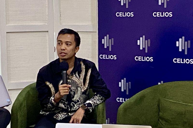 Seorang pria berbicara di podium dengan mikrofon sambil duduk di kursi hijau di depan latar belakang biru bertuliskan logo Celios.