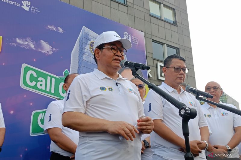 Seorang pria berkaos putih dan topi putih berbicara di depan mikrofon pada acara resmi di hadapan gedung berlantai banyak, dengan beberapa orang lain dalam kaos putih berdiri di sekitarnya.