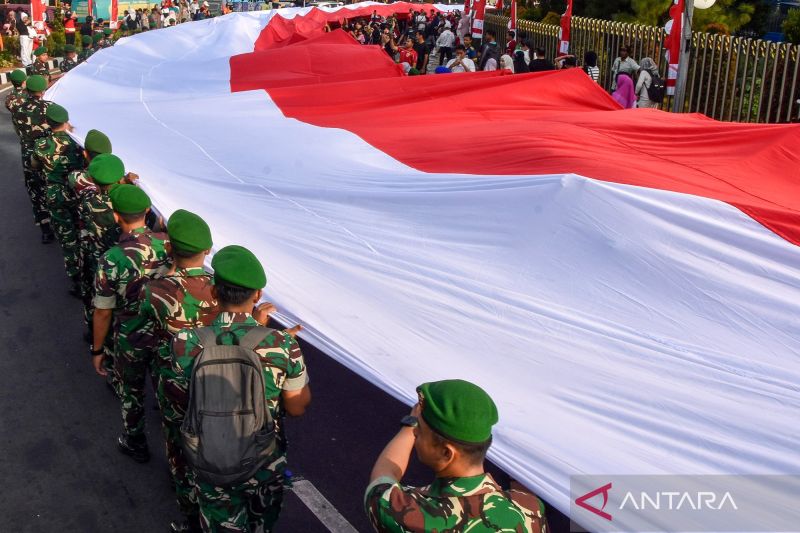 Deretan tentara dalam seragam hijau berderet di samping kain bendera Indonesia merah putih berukuran besar yang dikibarkan di jalan raya dengan penonton latar belakang.