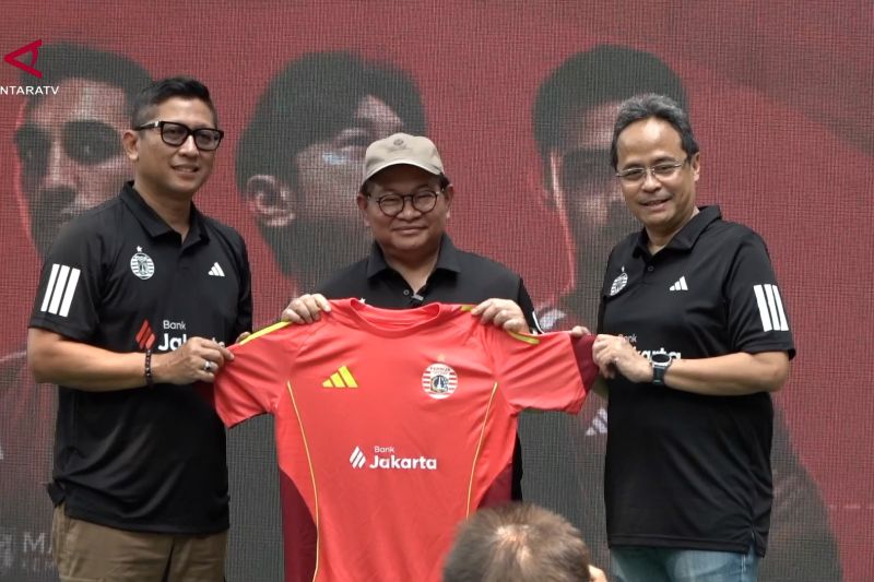 Tiga pria berbaris sambil memegang seragam olahraga merah dengan logo Adidas dan Bank Jakarta di depan layar LED merah.
