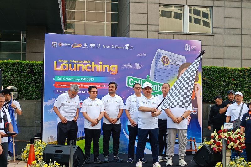 Para pejabat dan staf Transjakarta dalam pakaian putih berdiri di atas panggung di depan spanduk biru saat acara peluncuran rute Senayan-Ancol pada 8 Agustus 2023, dengan bunga dan bendera merah putih di sekitar mereka.