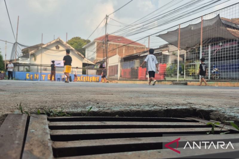 Lapangan bermain anak-anak dengan beberapa anak sedang bermain, dikelilingi bangunan rumah dan tangki penyimpanan air berwarna merah, dengan sistem saluran air dan pipa yang terlihat di permukaan tanah.