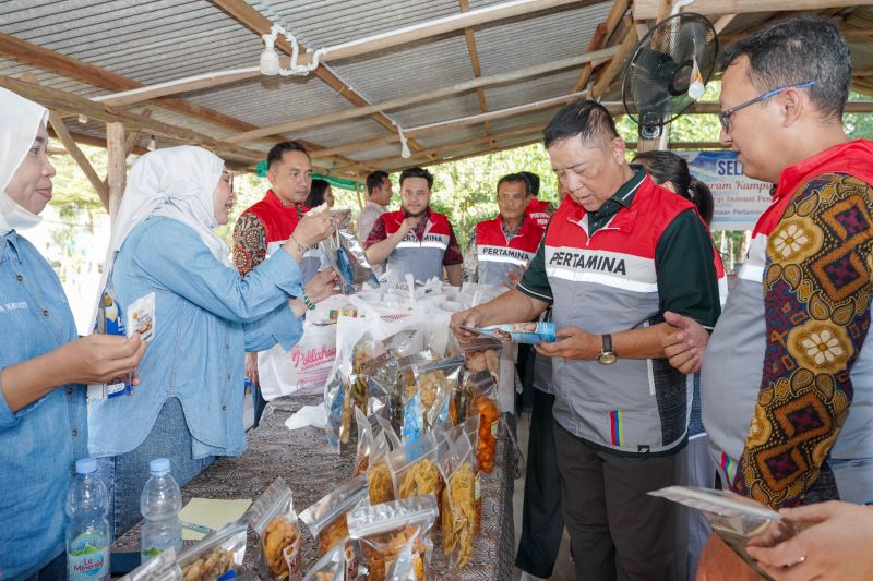 Sekelompok orang berkumpul di bawah tenda untuk melihat produk makanan laut olahan yang ditampilkan di meja, dengan beberapa orang mengenakan rompi merah bertuliskan Pertamina.